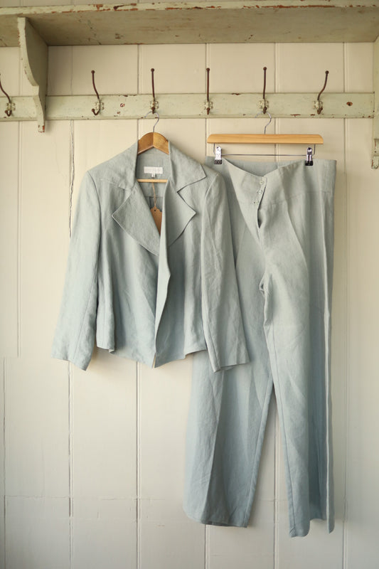  A sage green linen two-piece suit by Joyce Ridings, featuring a cropped jacket and matching high-waisted trousers, both hanging on a vintage wooden peg rail.
