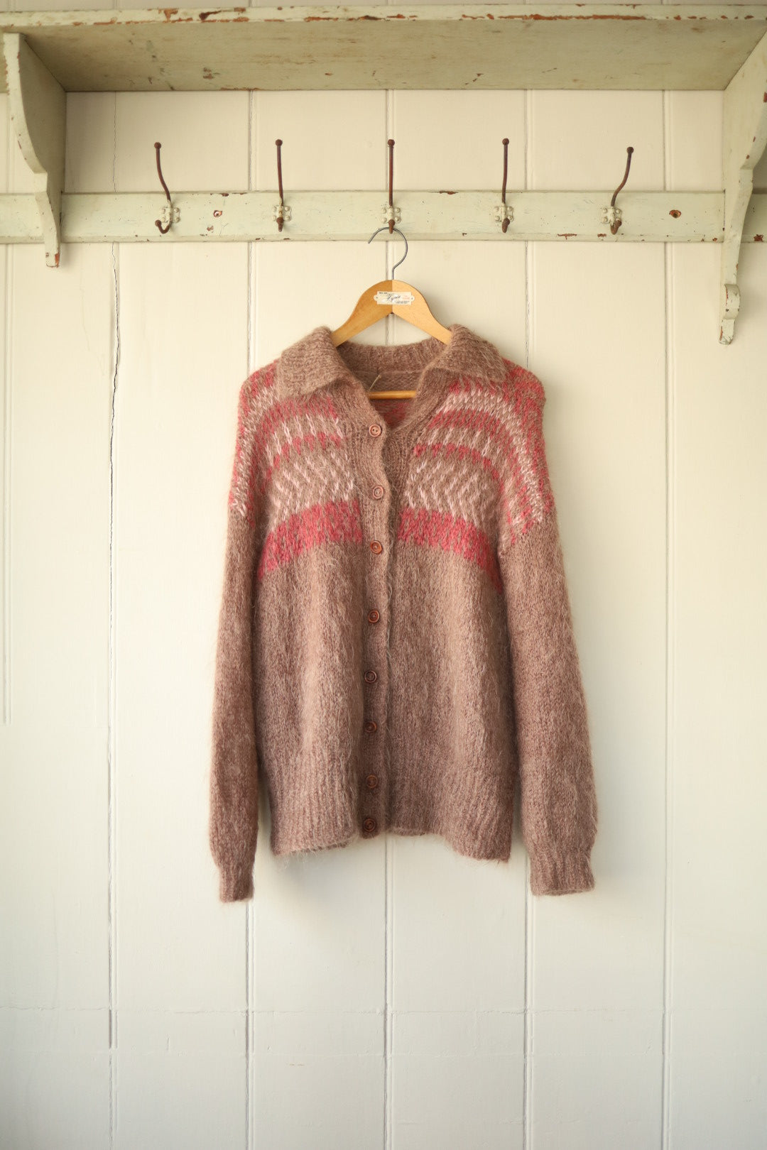 A handknitted mohair and wool cardigan with a slouchy fit, button front and intarsia pattern in warm pink and taupe tones, hanging on a wooden hanger against a panelled cream wall.
