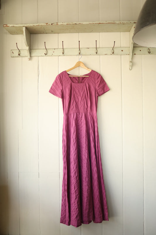 A mid-century style, home-sewn raspberry pink fit and flare dress with short sleeves and a square neckline, displayed on a wooden hanger against a pale panelled wall.
