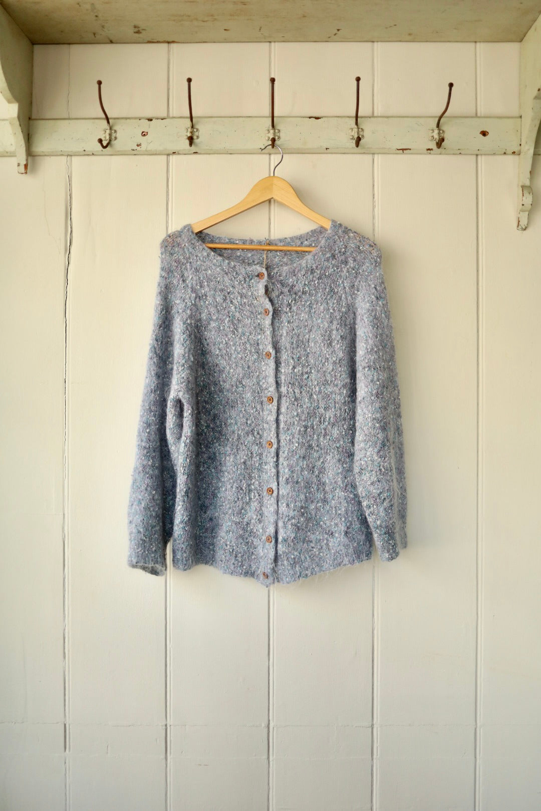 A hand-knitted flecked mohair cardigan in soft grey tones, displayed on a wooden hanger against a white panelled wall. The cardigan features a round neckline and wooden buttons down the front.