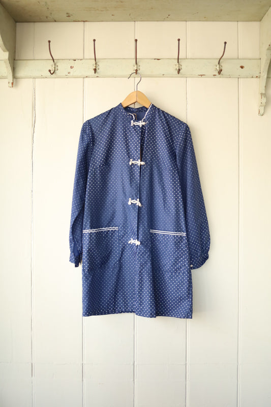 Buttoned jacket in navy with small white polka dots, mandarin collar, frog fastenings, and front patch pockets, displayed on a wooden hanger against a panelled wall.