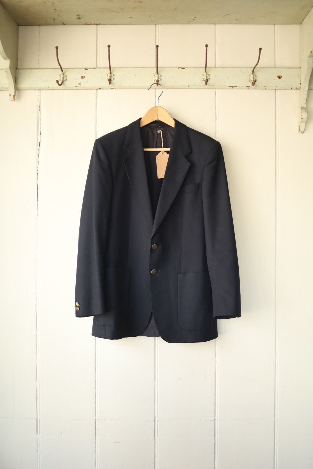Classic navy wool blazer with two-button fastening and patch pockets, hanging on a wooden hanger against a painted white panelled wall.