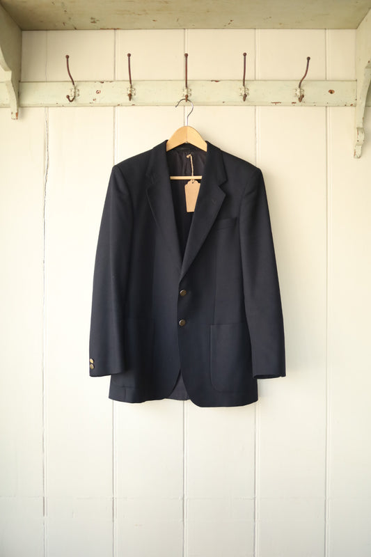 Classic navy wool blazer with two-button fastening and patch pockets, hanging on a wooden hanger against a painted white panelled wall.