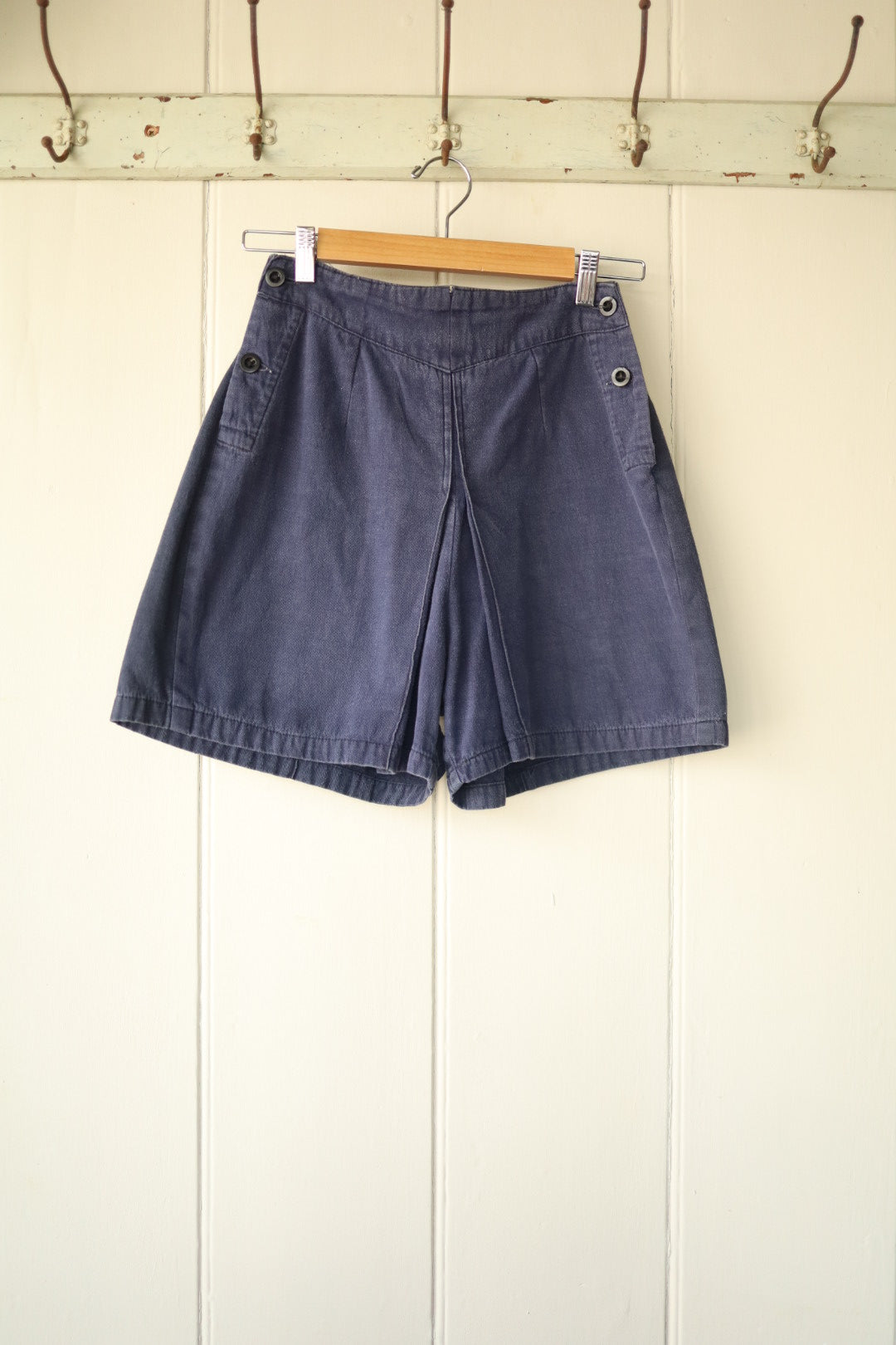 Front view of vintage 1940s navy blue workwear shorts with wide leg fit and side button detailing, hanging on a wooden hanger against a cream panelled wall.