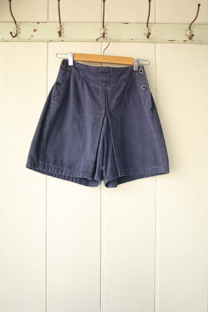 Front view of vintage 1940s navy blue workwear shorts with wide leg fit and side button detailing, hanging on a wooden hanger against a cream panelled wall.