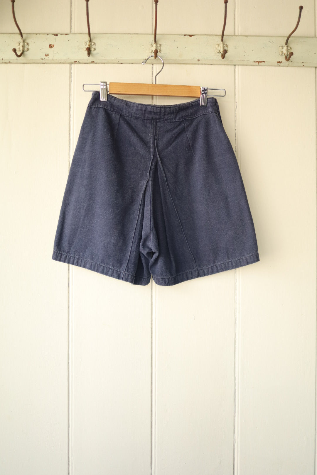 Rear view of the same navy blue vintage workwear shorts, showing tailored seam lines and simple waistband design, hung on a wooden hanger.
