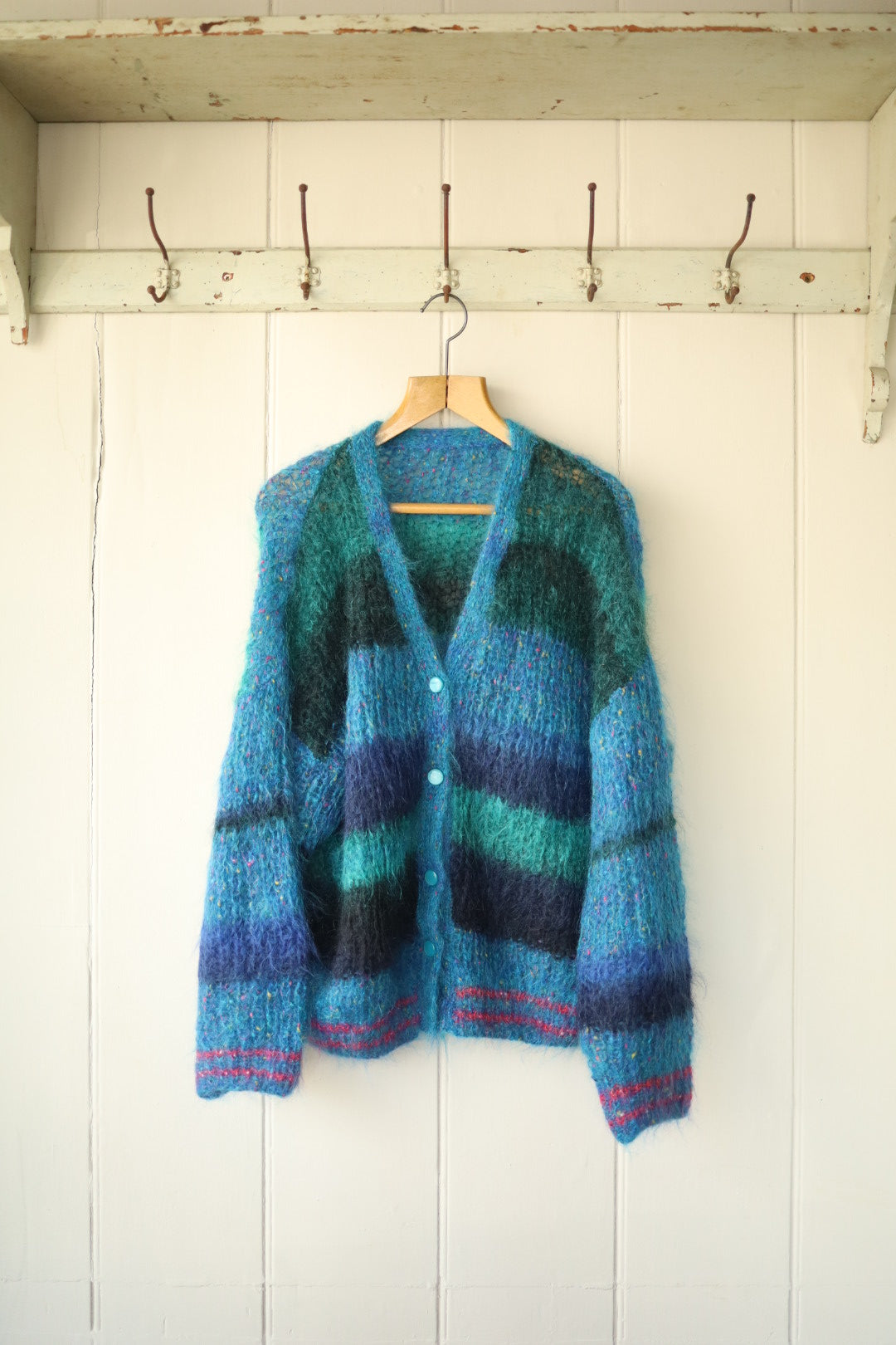 Front view of a hand-knitted mohair cardigan in wide horizontal stripes of teal, navy, and turquoise, with a V-neckline, long sleeves, and blue buttons down the centre. The cardigan hangs on a wooden hanger against a painted wooden wall.”