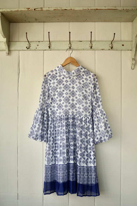 A 1970s-style white cheesecloth dress with a blue geometric print, featuring dramatic balloon sleeves, a high collar, and a gathered waist, displayed hanging against a panelled wall.