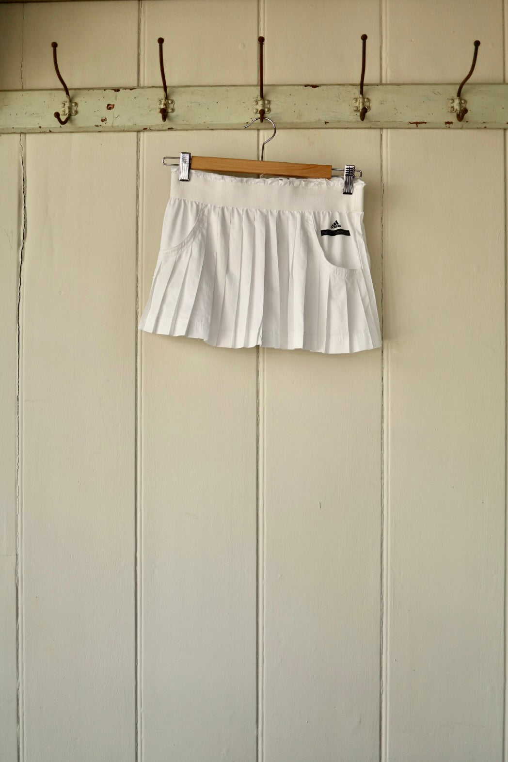 A white Stella McCartney pleated tennis skirt, hanging on a wooden hanger against a panelled wall with hooks.