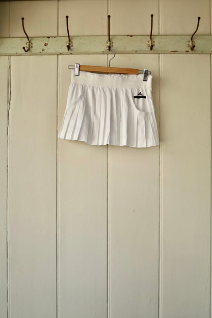 A white Stella McCartney pleated tennis skirt, hanging on a wooden hanger against a panelled wall with hooks.