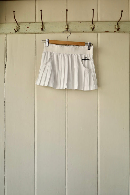A white Stella McCartney pleated tennis skirt, hanging on a wooden hanger against a panelled wall with hooks.