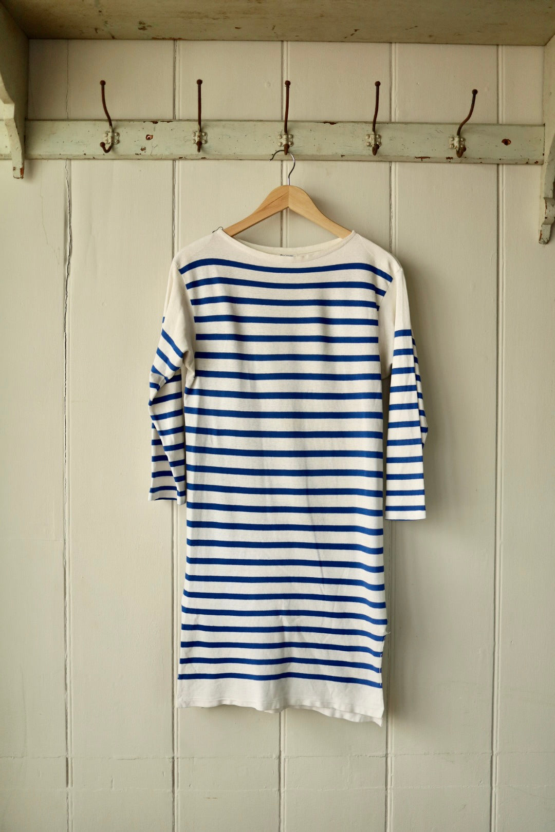 Back view of a vintage Orcival Breton dress in cream with classic cobalt blue stripes, displayed on a wooden hanger against a painted wooden wall with iron hooks.