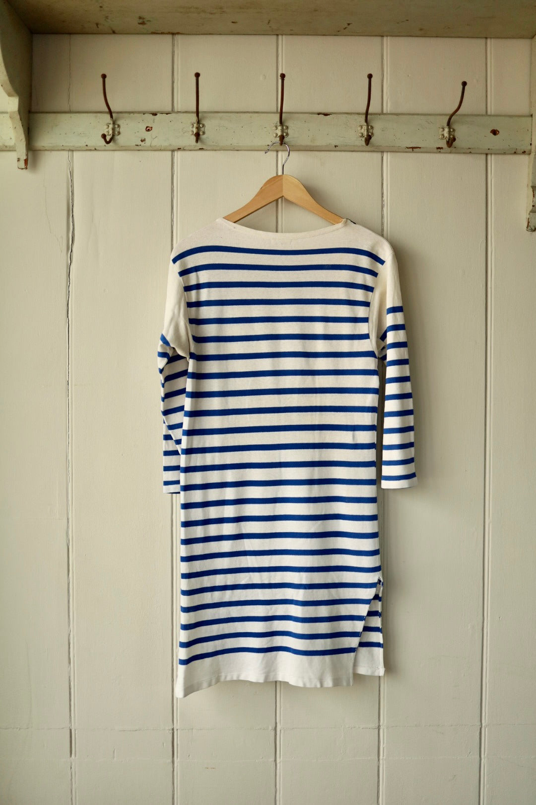 Rear view of an Orcival Breton dress in cream with cobalt blue horizontal stripes and side vent detail, displayed on a wooden hanger against a pale wooden panel wall with vintage iron hooks.
