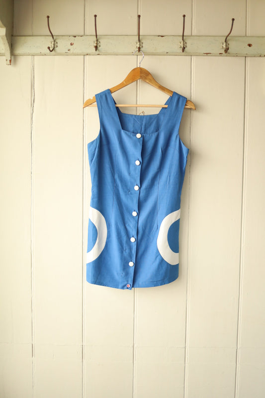 Front view of a vintage 1960s sleeveless blue mini dress with bold white circle appliqués on the hips and white buttons down the front, displayed on a wooden hanger against a cream panelled wall with iron hooks.