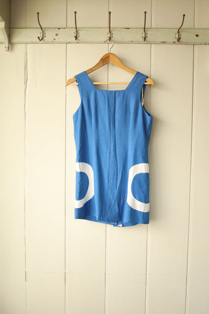 Back view of a vintage 1960s Mod-style sleeveless mini dress in cobalt blue with bold white semi-circle appliqués on the hips, displayed on a wooden hanger against a cream wooden panel wall.
