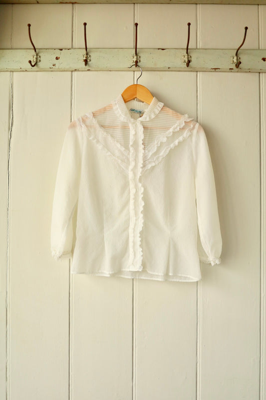 Front view of a vintage white sheer blouse with ruffled lace trim, high neckline, and long sleeves, displayed on a wooden hanger against a pale panelled wall with antique iron hooks.