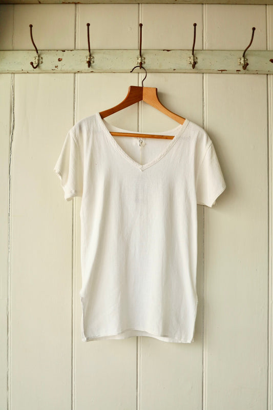 Front view of a vintage 1960s white Airtex top with a V-neckline and short sleeves, made from lightweight textured cotton, displayed on a wooden hanger against a pale panelled wall with antique iron hooks.