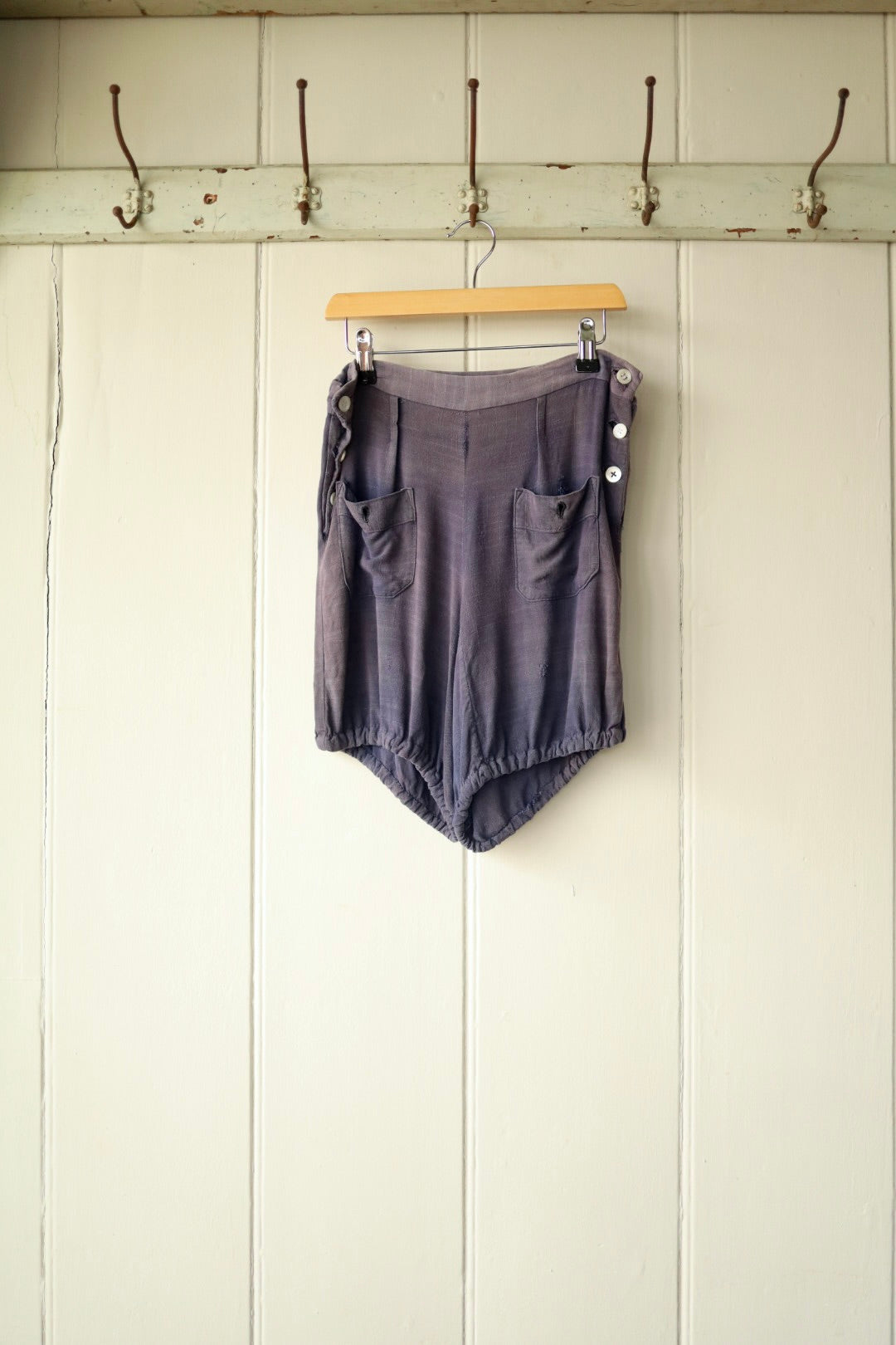 Vintage 1930s women's workwear shorts in soft faded blue cotton-linen fabric, with high waist, side button fastening, front patch pockets, and gathered leg hems, displayed on a wooden hanger against a cream panelled wall with iron hooks.