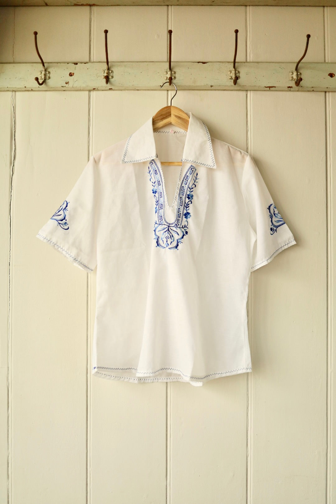 Front view of a vintage white short-sleeved shirt with blue embroidery at the neckline and sleeves, featuring a pointed collar and decorative stitch trim, displayed on a wooden hanger against a cream panelled wall.
