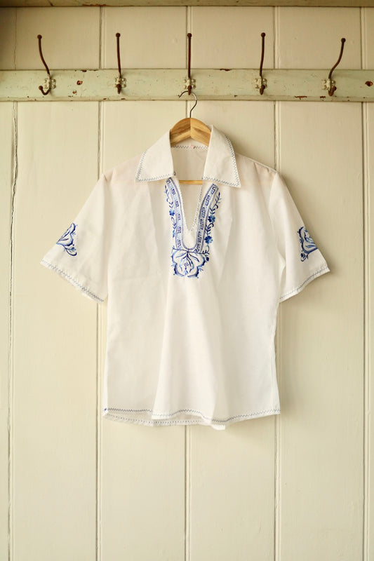 Front view of a vintage white short-sleeved shirt with blue embroidery at the neckline and sleeves, featuring a pointed collar and decorative stitch trim, displayed on a wooden hanger against a cream panelled wall.