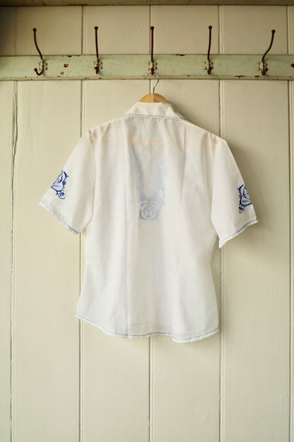 Back view of the vintage white embroidered shirt, showing the semi-sheer cotton fabric and subtle reverse of the blue front embroidery, displayed on a wooden hanger against a cream panelled wall.