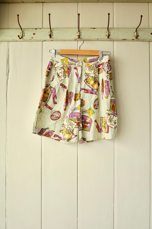 Front view of vintage printed shorts with a novelty travel-themed print in purple, yellow, red, and green tones on a pale patterned background, displayed on a hanger against a cream panelled wall.