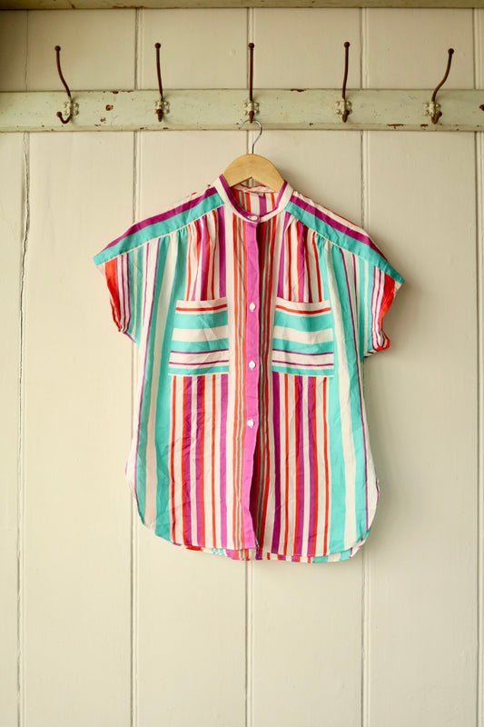 Front view of a vibrant vintage blouse with vertical stripes in pink, red, aqua, and purple tones, featuring short sleeves, white buttons, and two contrasting striped patch pockets, displayed on a wooden hanger against a cream panelled wall.