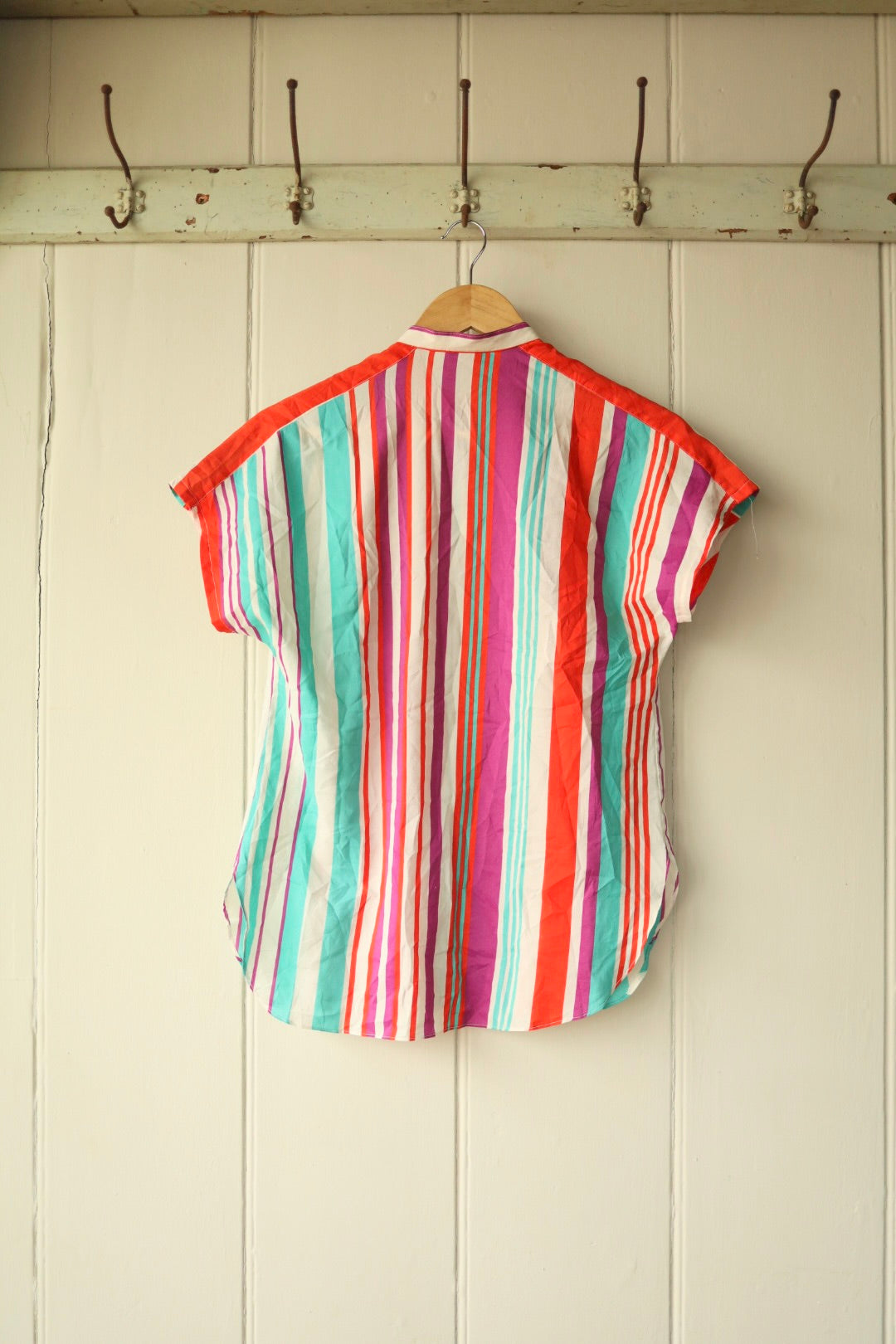 Back view of the same vintage striped blouse, showing colourful vertical stripes in bright tones with a curved hem and short sleeves, hanging against a cream panelled wall with antique hooks.