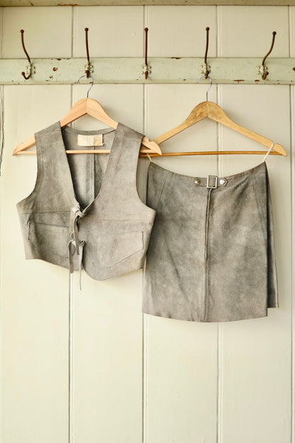 Back view of a vintage 1970s grey suede two-piece set, featuring a cropped sleeveless vest and A-line mini skirt, both hanging on wooden hangers against a painted wooden wall with hooks.