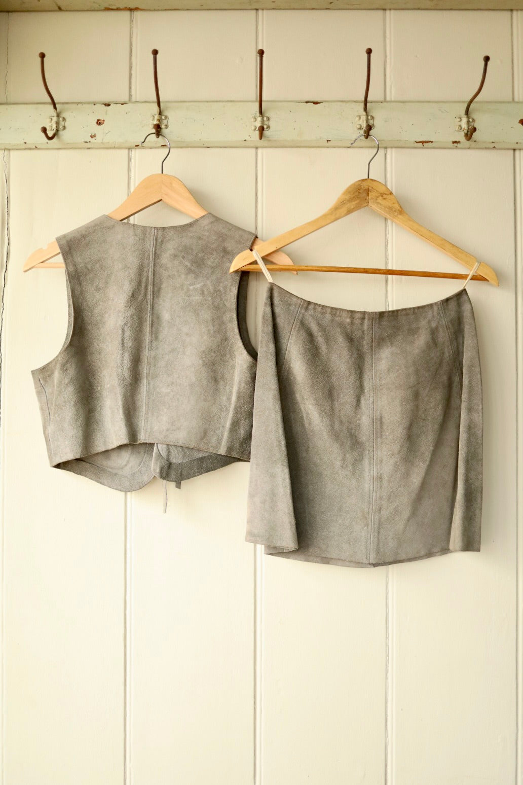 Front view of a vintage 1970s grey suede co-ord set showing the cropped vest with tie front and patch pockets, and the matching A-line mini skirt with zip, buckle, and press stud details, displayed on wooden hangers.