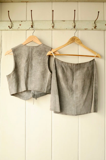 Front view of a vintage 1970s grey suede co-ord set showing the cropped vest with tie front and patch pockets, and the matching A-line mini skirt with zip, buckle, and press stud details, displayed on wooden hangers.