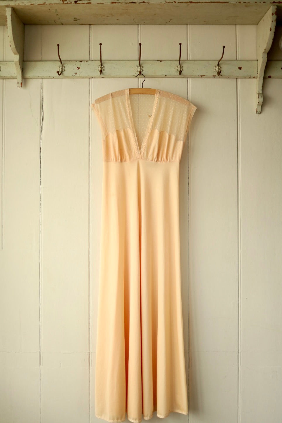 Front view of a vintage peach nightdress with a deep V neckline and delicate polka dot mesh bodice, hanging from a wooden hanger against a cream-painted wooden wall