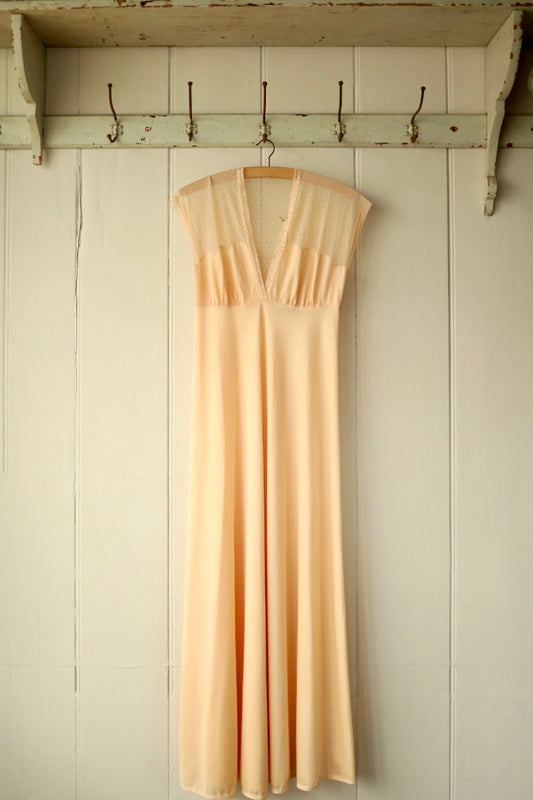 Front view of a vintage peach nightdress with a deep V neckline and delicate polka dot mesh bodice, hanging from a wooden hanger against a cream-painted wooden wall