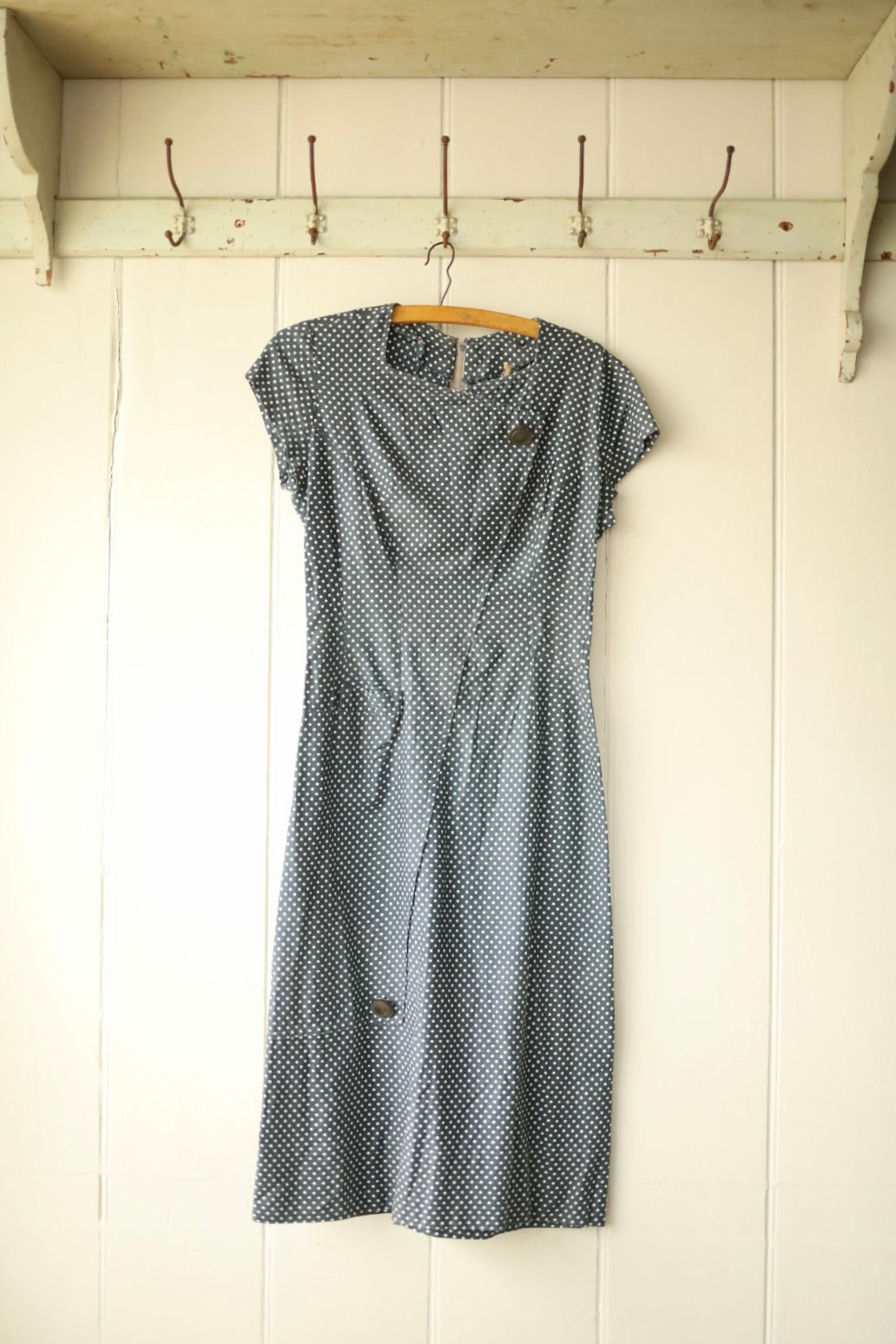 Front view of a vintage navy blue dress with short sleeves and white polka dots, featuring an asymmetric button detail at the neckline and soft pleats at the waist, hanging on a wooden hanger against a light wooden wall