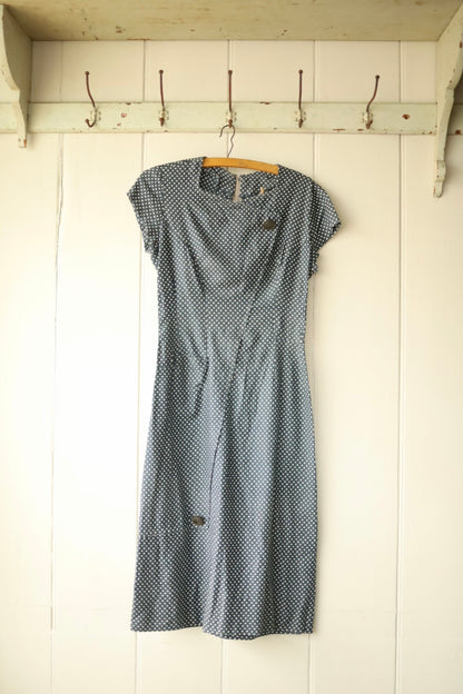 Front view of a vintage navy blue dress with short sleeves and white polka dots, featuring an asymmetric button detail at the neckline and soft pleats at the waist, hanging on a wooden hanger against a light wooden wall