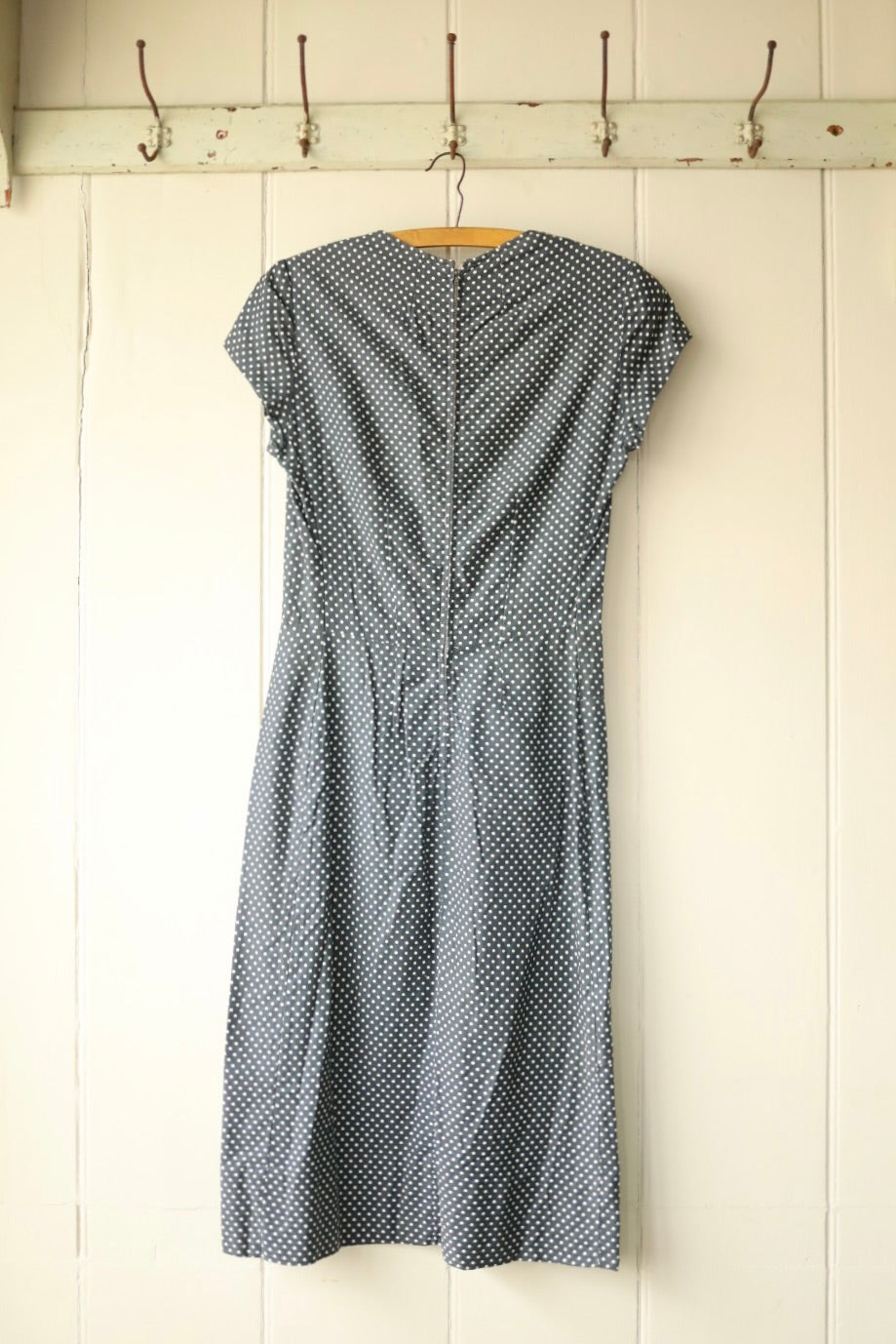 Back view of a vintage navy blue polka dot dress with a fitted waist and zip closure, displayed on a wooden hanger against a pale wooden panelled wall.