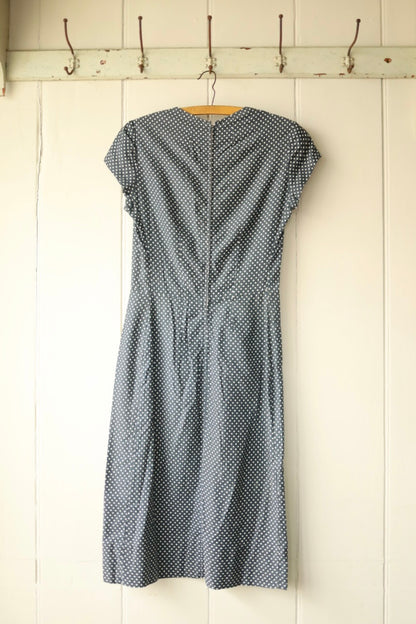 Back view of a vintage navy blue polka dot dress with a fitted waist and zip closure, displayed on a wooden hanger against a pale wooden panelled wall.
