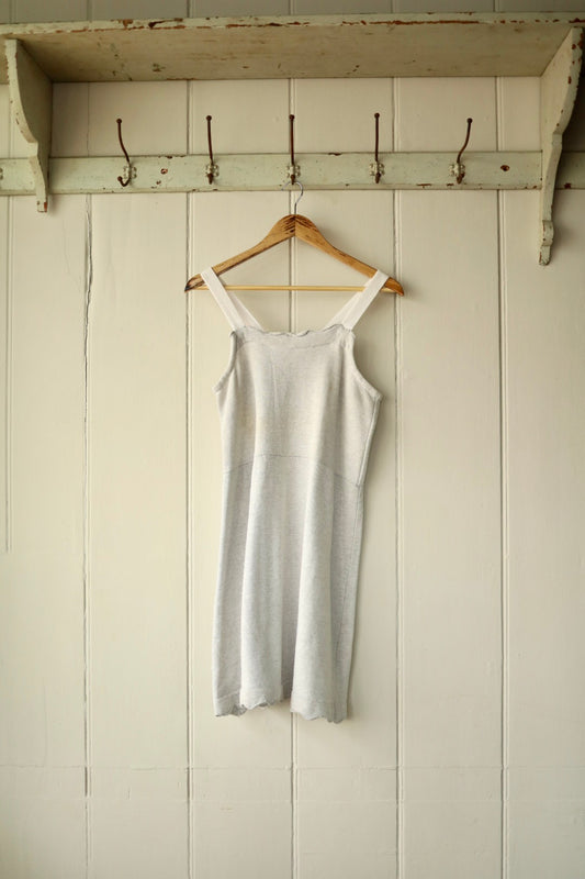 Front view of an antique French sleeveless undergarment dress in marl grey jersey with thick straps and a scalloped hem, displayed on a wooden hanger against a cream panelled wall.