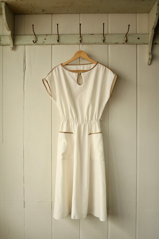 Front view of a vintage cream day dress featuring brown contrast trim, patch pockets, a gathered waist, and a keyhole neckline detail, displayed on a wooden hanger against a cream-painted wooden wall