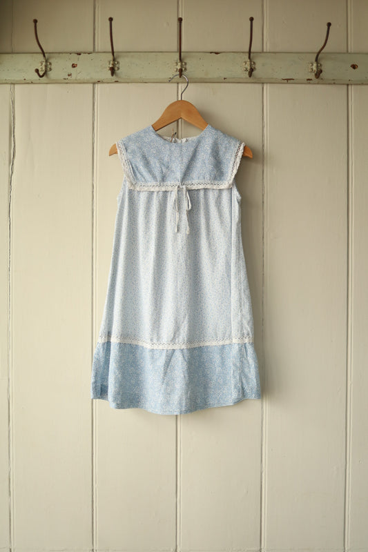 Front view of a sleeveless vintage ditsy floral dress in soft blue tones with bib-style yoke, lace trim, and tie detail, displayed on a wooden hanger against a painted wooden wall.