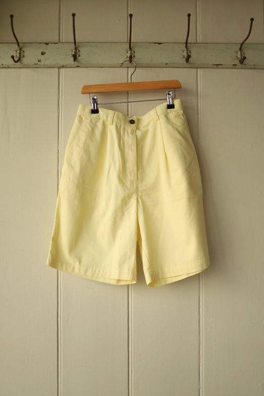 Front view of vintage lemon yellow pleated shorts with side pockets, belt loops, and single button fastening, hanging on a wooden hanger against a cream panelled wall.