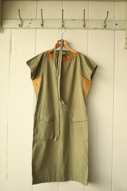 Front view of a two-tone khaki dress with cap sleeves, tan contrast yoke and underarm panels, patch pockets, and tie at waist, hanging on a wooden hanger against a cream wall.