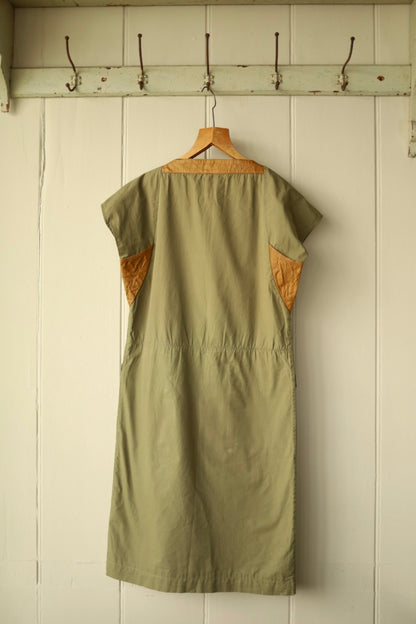 Back view of the khaki dress with tan yoke and underarm inserts, shown against a cream painted wooden wall.