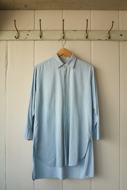 Front view of oversized pale blue striped shirt hanging against a vintage cream panelled wall with hooks above.
