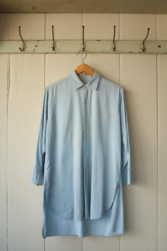Front view of oversized pale blue striped shirt hanging against a vintage cream panelled wall with hooks above.
