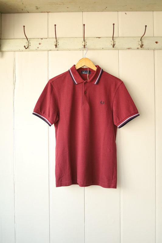 Front view of a Fred Perry maroon cotton polo shirt with navy and white tipping on the collar and sleeves, hanging on a wooden hanger against a cream tongue-and-groove wall with vintage metal hooks above.