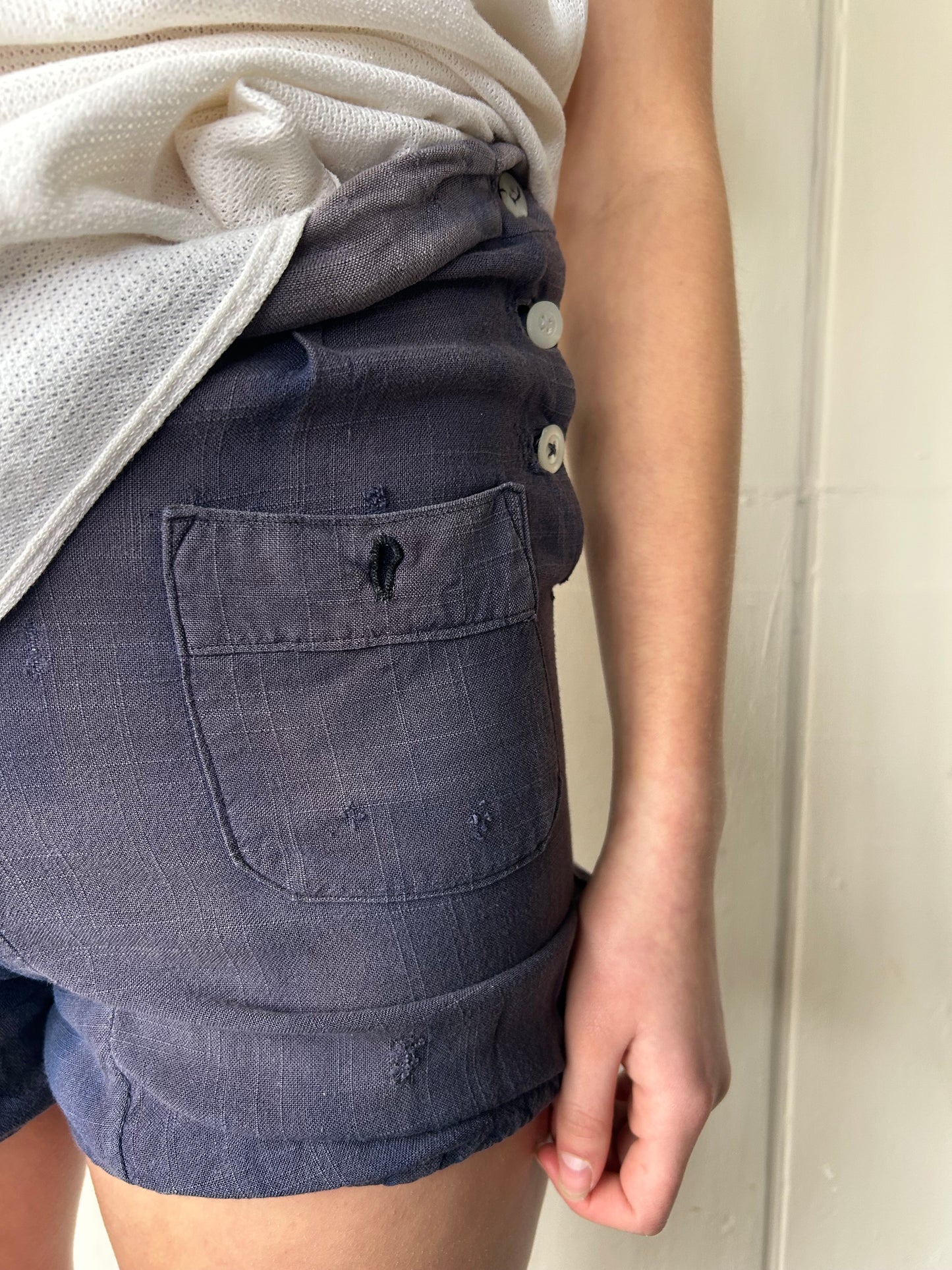 Close-up of a model wearing vintage 1930s faded blue workwear shorts with patch pocket, side button fastening, and visible mended repairs, paired with a tucked cream textured top.