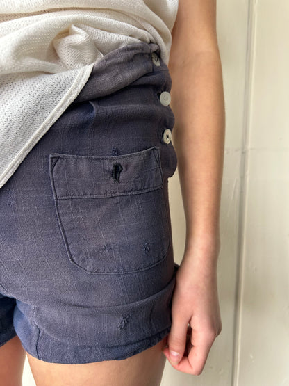 Close-up of a model wearing vintage 1930s faded blue workwear shorts with patch pocket, side button fastening, and visible mended repairs, paired with a tucked cream textured top.