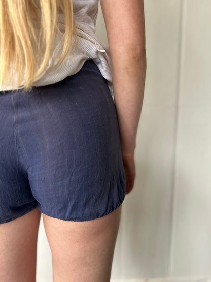 Rear close-up of a model wearing 1930s faded blue workwear shorts with gathered hems, showing the soft texture and drape of the cotton-linen fabric.
